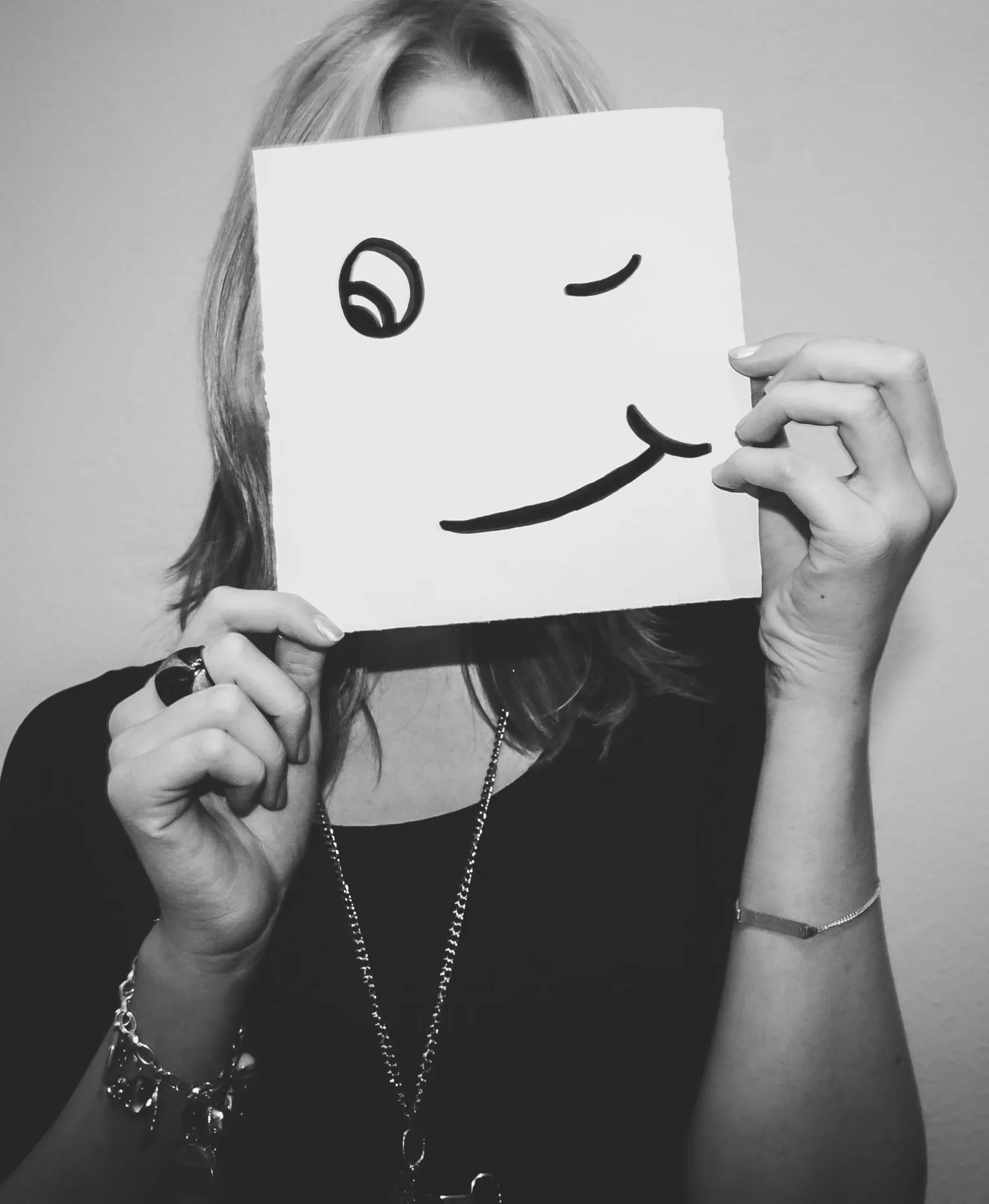 A black-and-white photo of a woman hiding her face behind a sheet of paper with a hand-drawn winking smiley face
