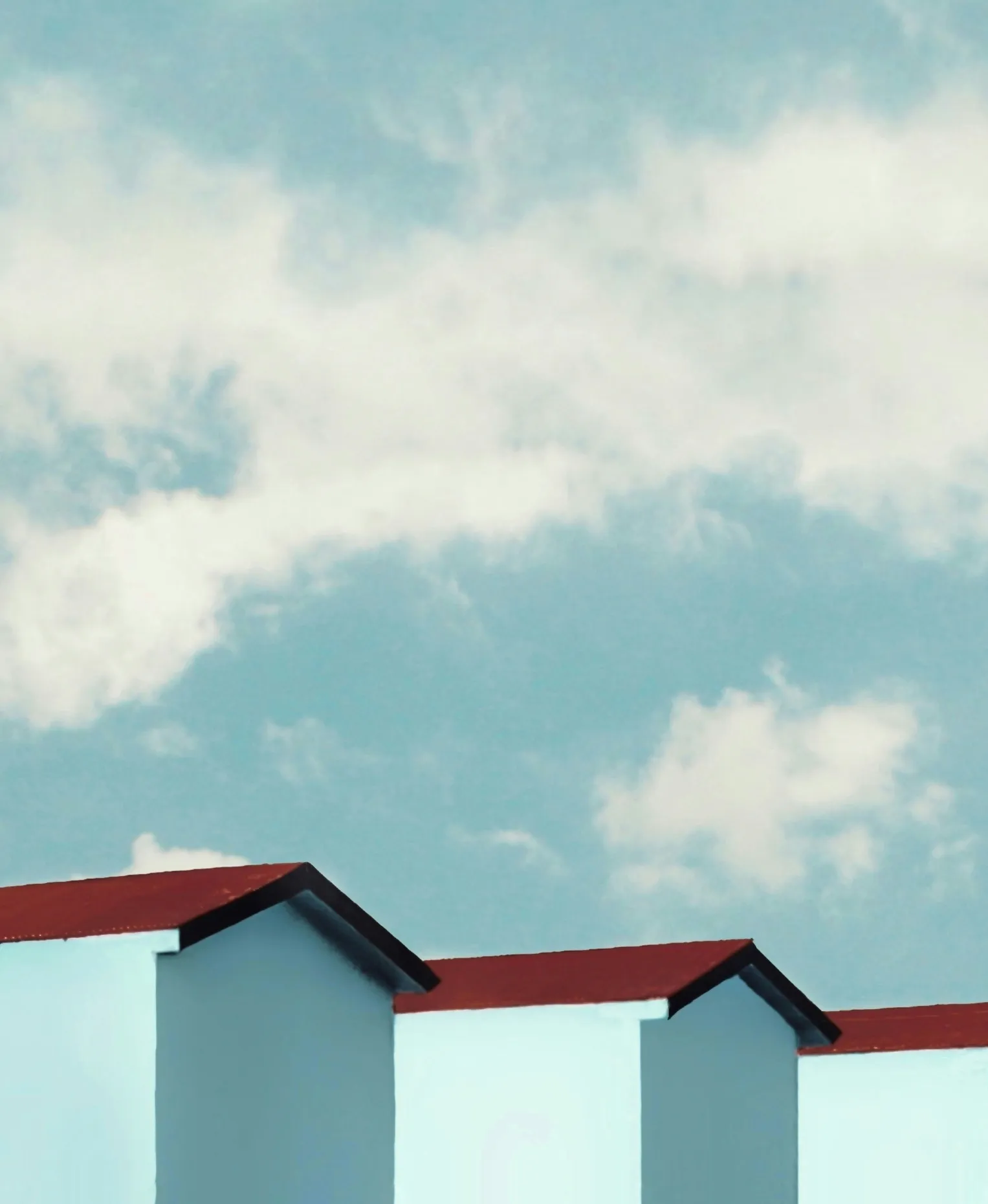 Three pale blue gabled buildings with red roofs in a row beneath a blue sky with drifting white clouds