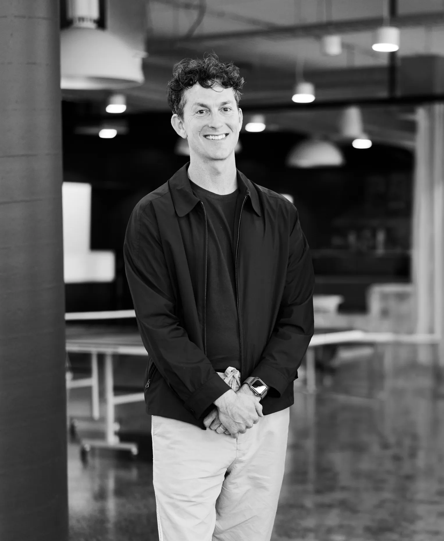 A black-and-white portrait of a smiling man with curly hair in a dark jacket, hands clasped, standing in an open-plan office