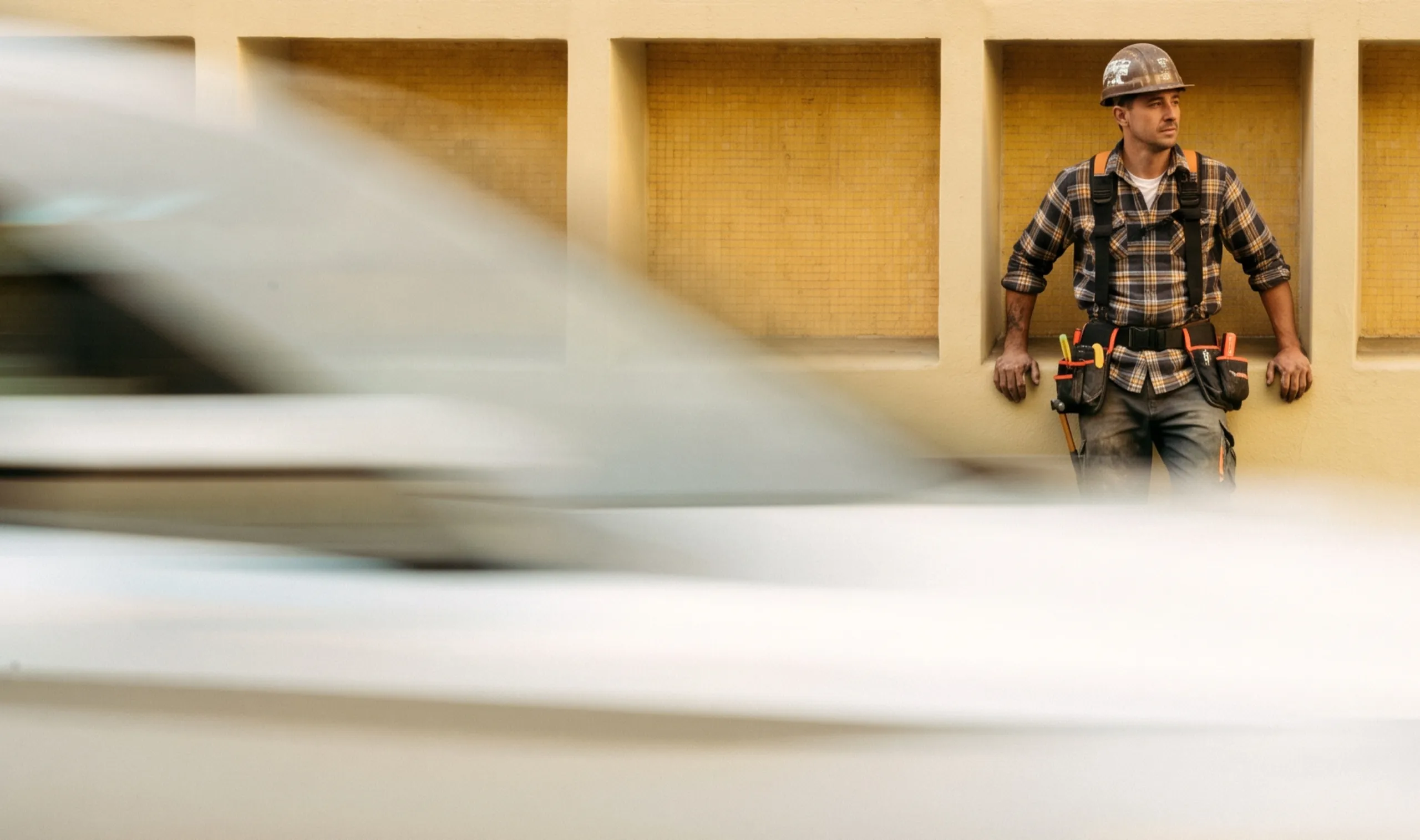 A tradie in a hard hat and tool belt leaning against a timber-panelled wall as a white van speeds past in a blur