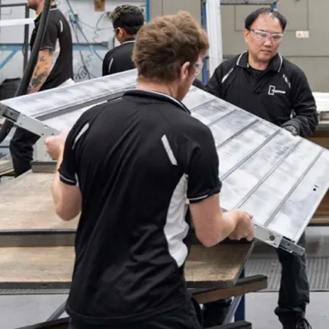 Two Parkwood factory workers in black polos carrying a large flat panel through the workshop
