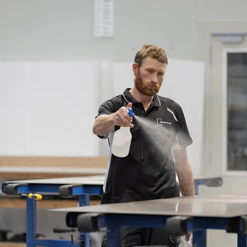 A bearded worker in a black polo spraying a bottle over a workbench in the factory