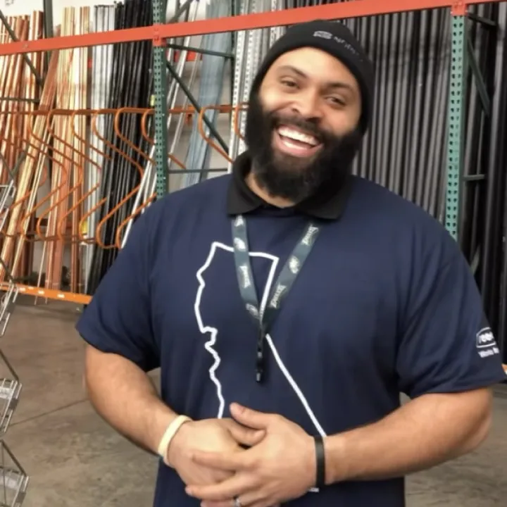 A smiling bearded Reece team member in a navy polo, beanie and lanyard, standing in a warehouse