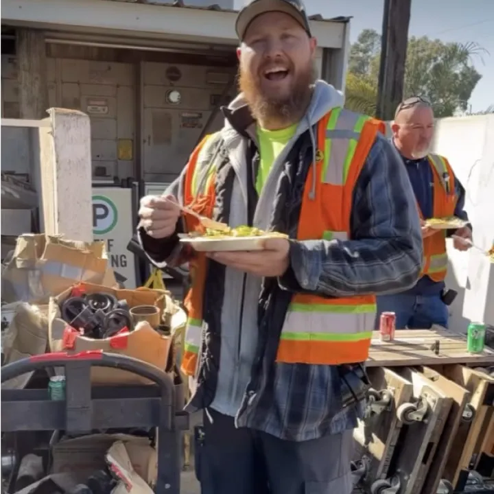 A laughing tradesman in a cap and orange hi-vis vest holding a plate of food at an outdoor yard event