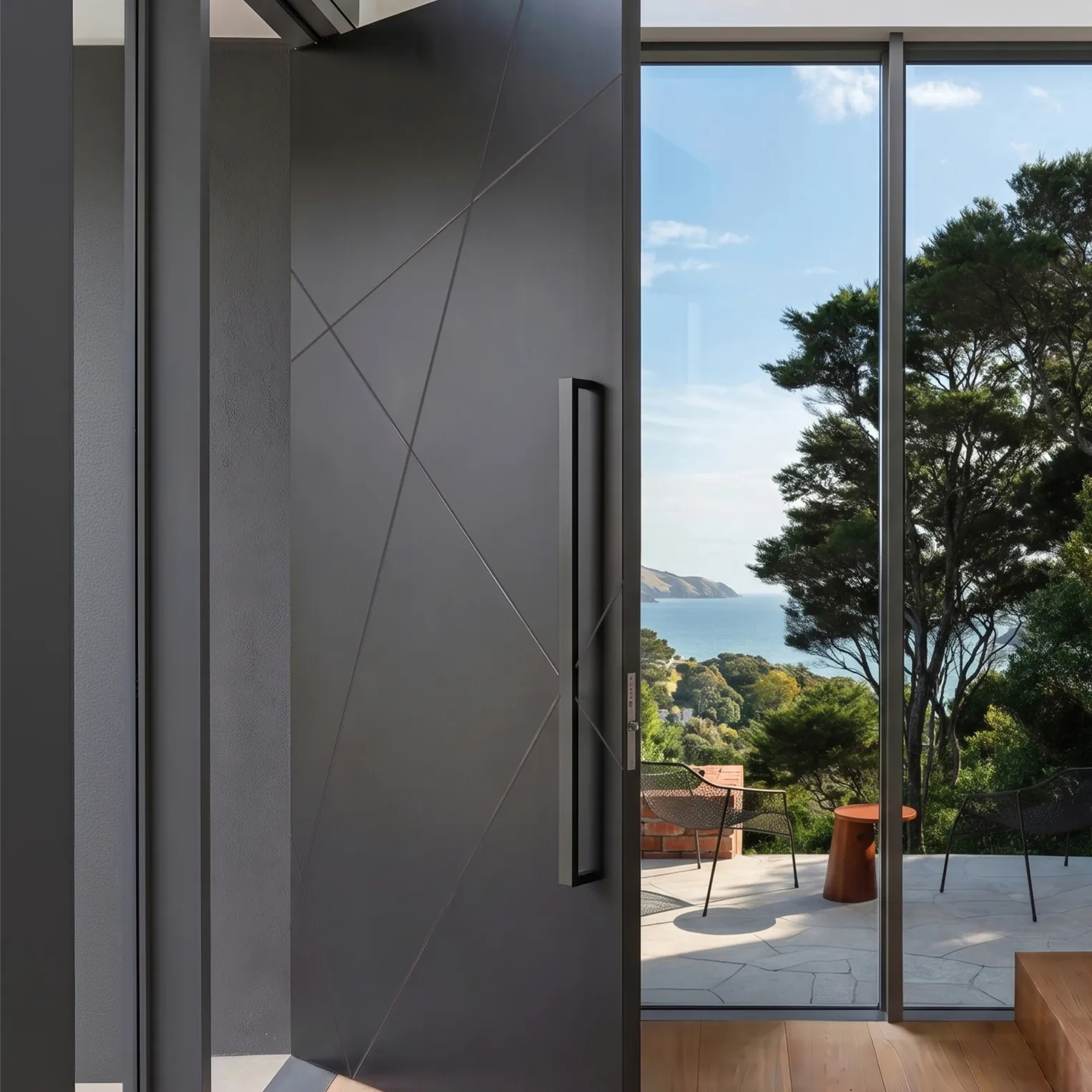 A charcoal pivot door with angled grooves and a long bar handle, opening onto a terrace with sea views