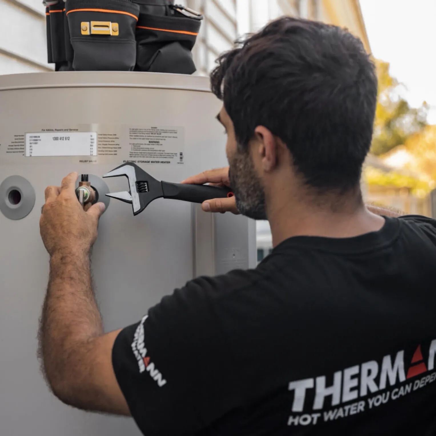A tradie in a black Thermann T-shirt tightening a fitting on a hot water tank, “Thermann Hot water you can depend on” printed across his back
