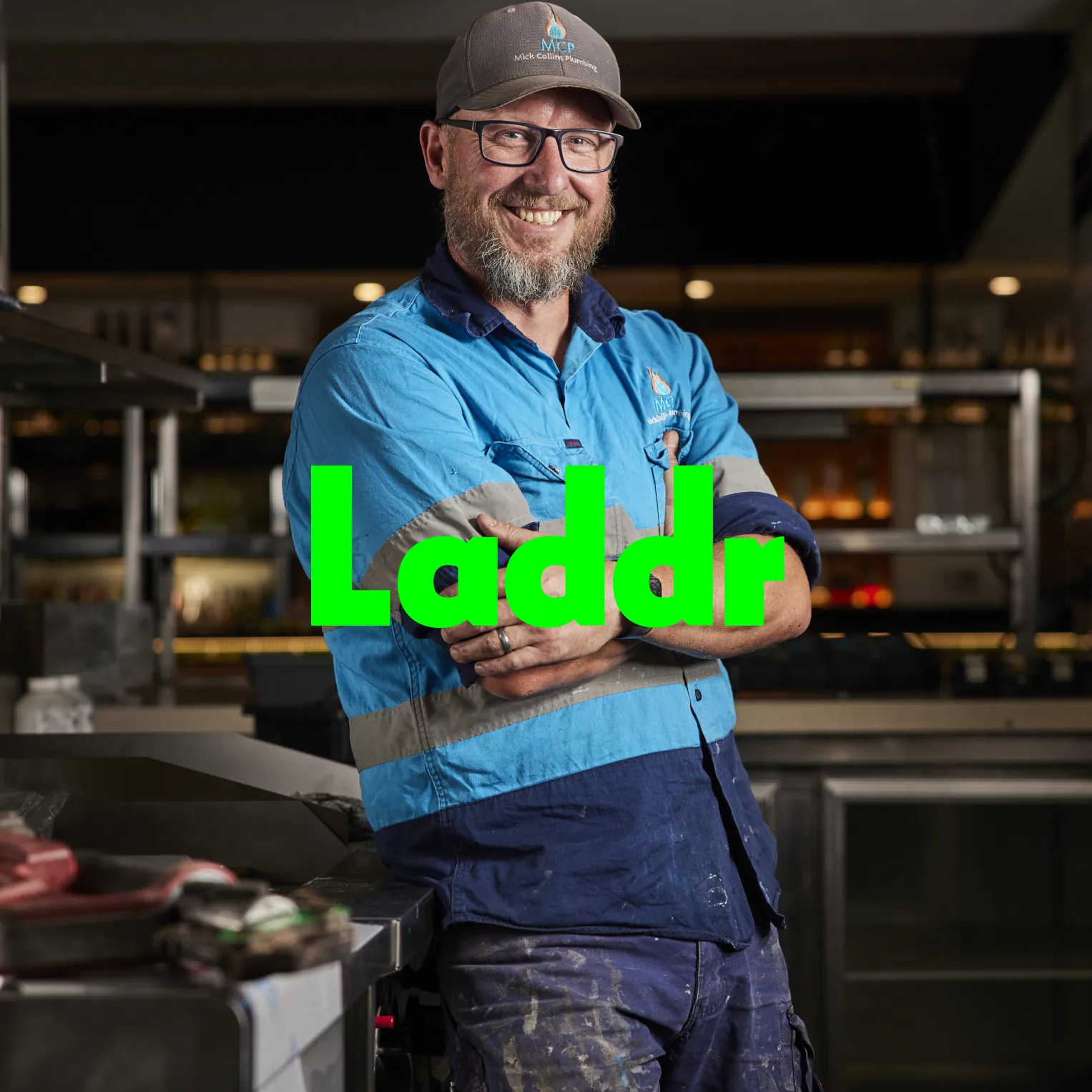 Laddr: a grinning plumber in a cap and blue work shirt with arms folded in a commercial kitchen, the Laddr logo in large green letters across the photo