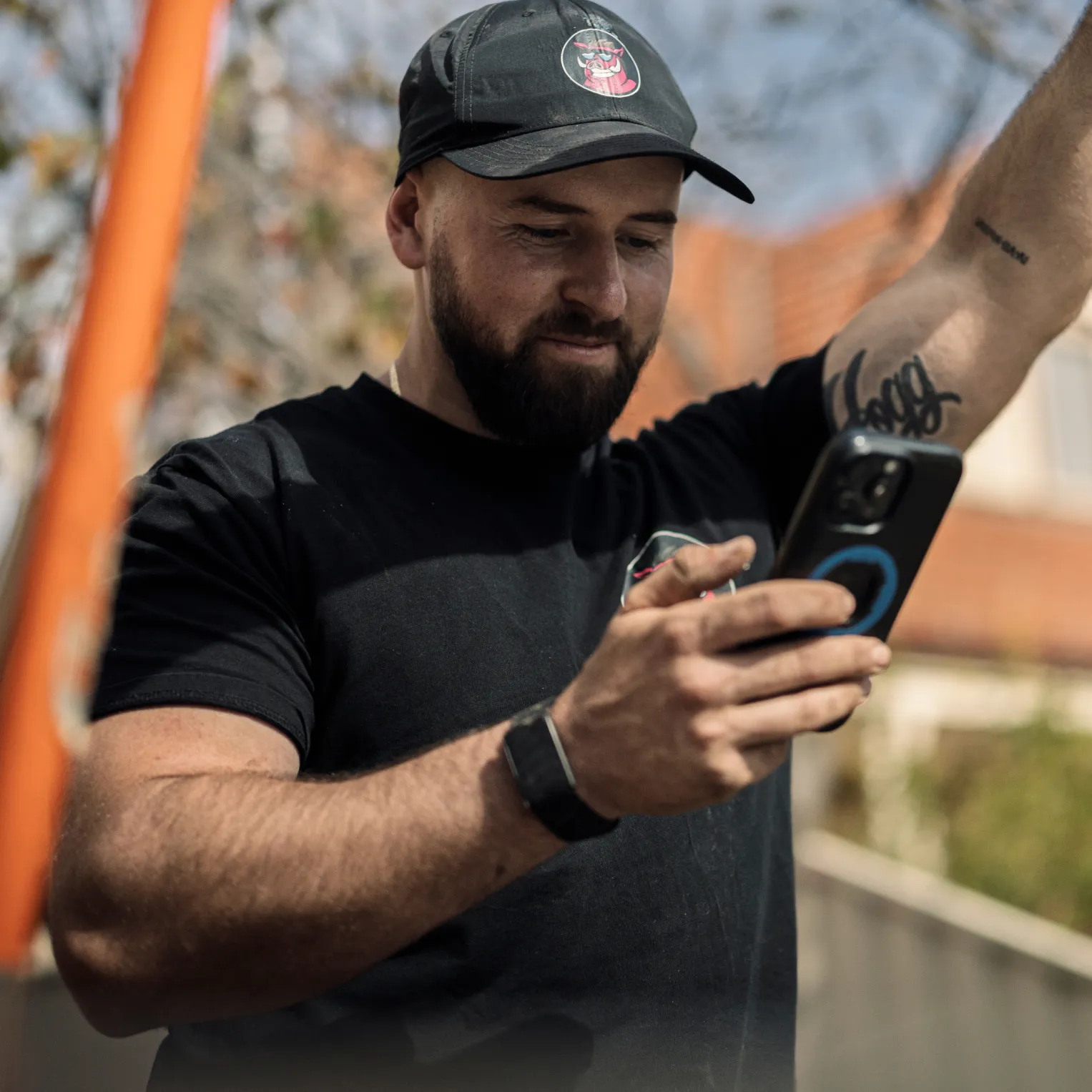 A bearded tradie in a black cap and T-shirt smiling down at his phone outdoors