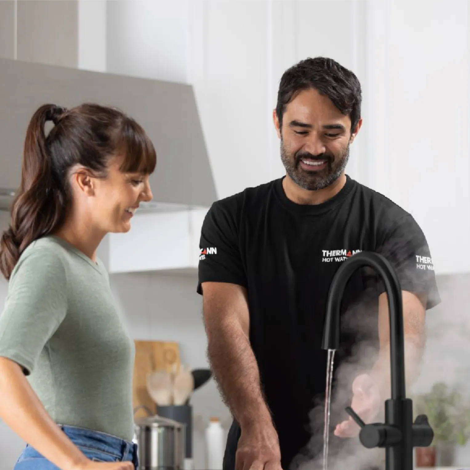 A smiling tradie in a black Thermann T-shirt running steaming water from a black kitchen tap while a woman looks on