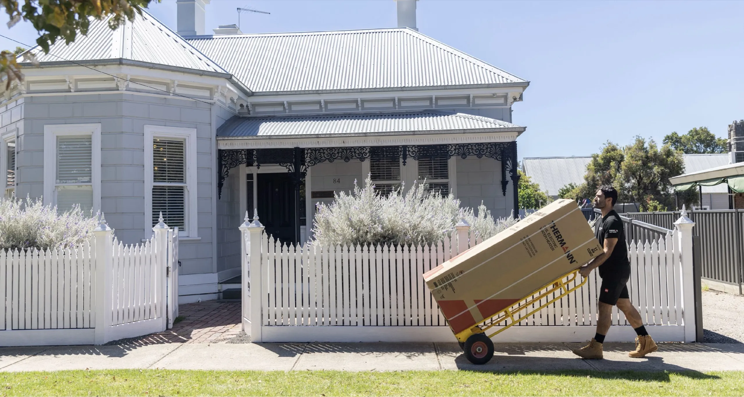 A tradie in a black Thermann T-shirt wheeling a boxed Thermann hot water unit on a yellow hand trolley past the white picket fence of a weatherboard house