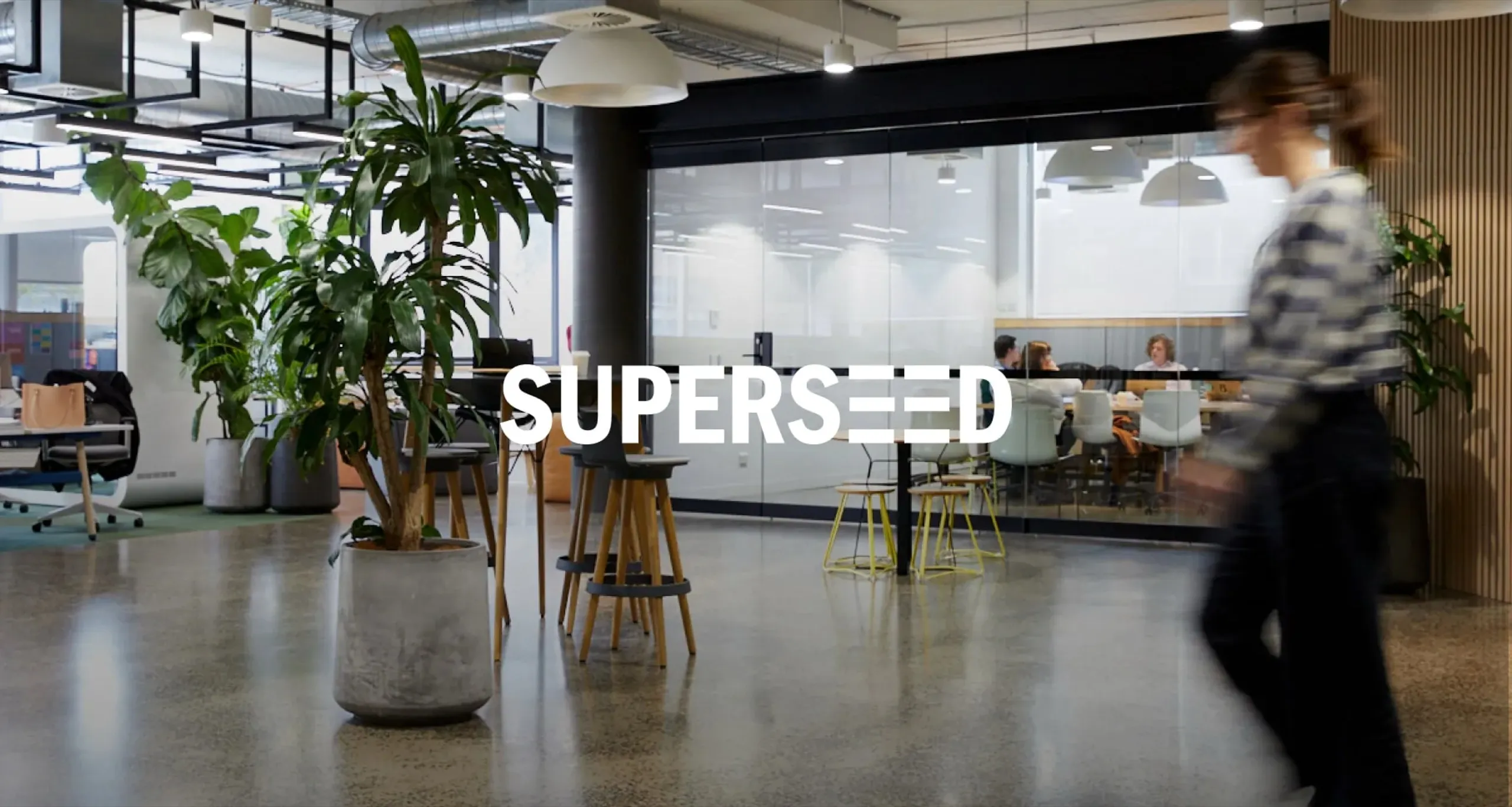 Superseed: an open-plan office with potted plants and a glass-walled meeting room, a woman blurred in motion as she walks past, the Superseed wordmark across the centre