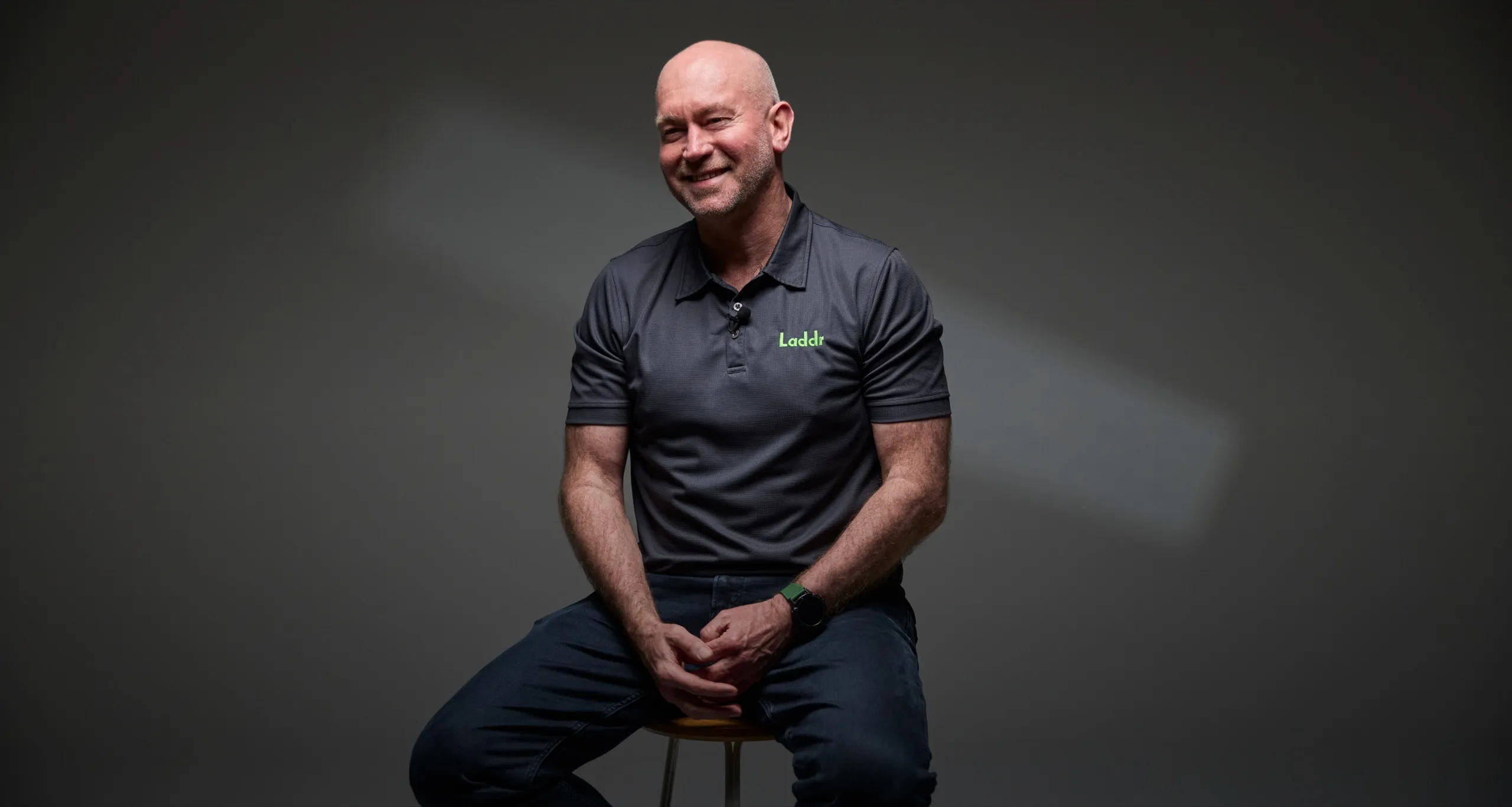 Laddr CEO Andrew Watt smiling on a stool against a dark grey backdrop, in a charcoal polo with a green Laddr logo