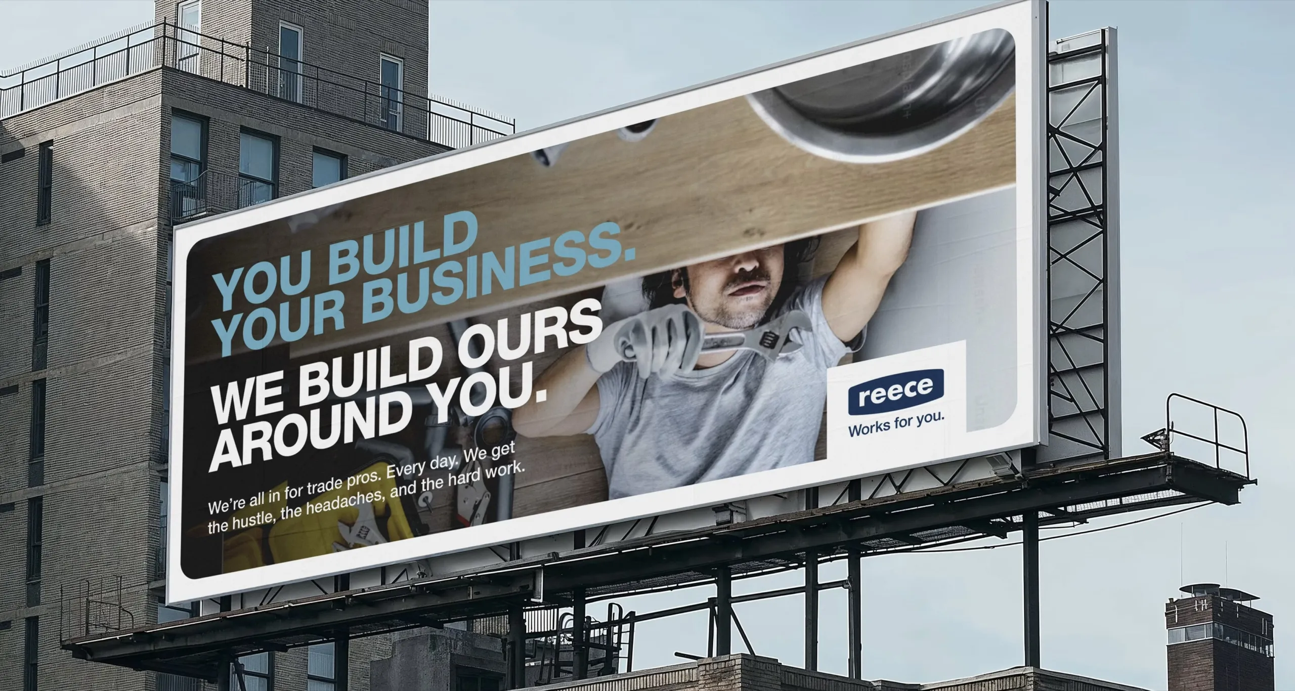 A Reece billboard above a city street reading “You build your business. We build ours around you.” beside the Reece logo and “Works for you.”, with a tradesman lying on his back holding a wrench