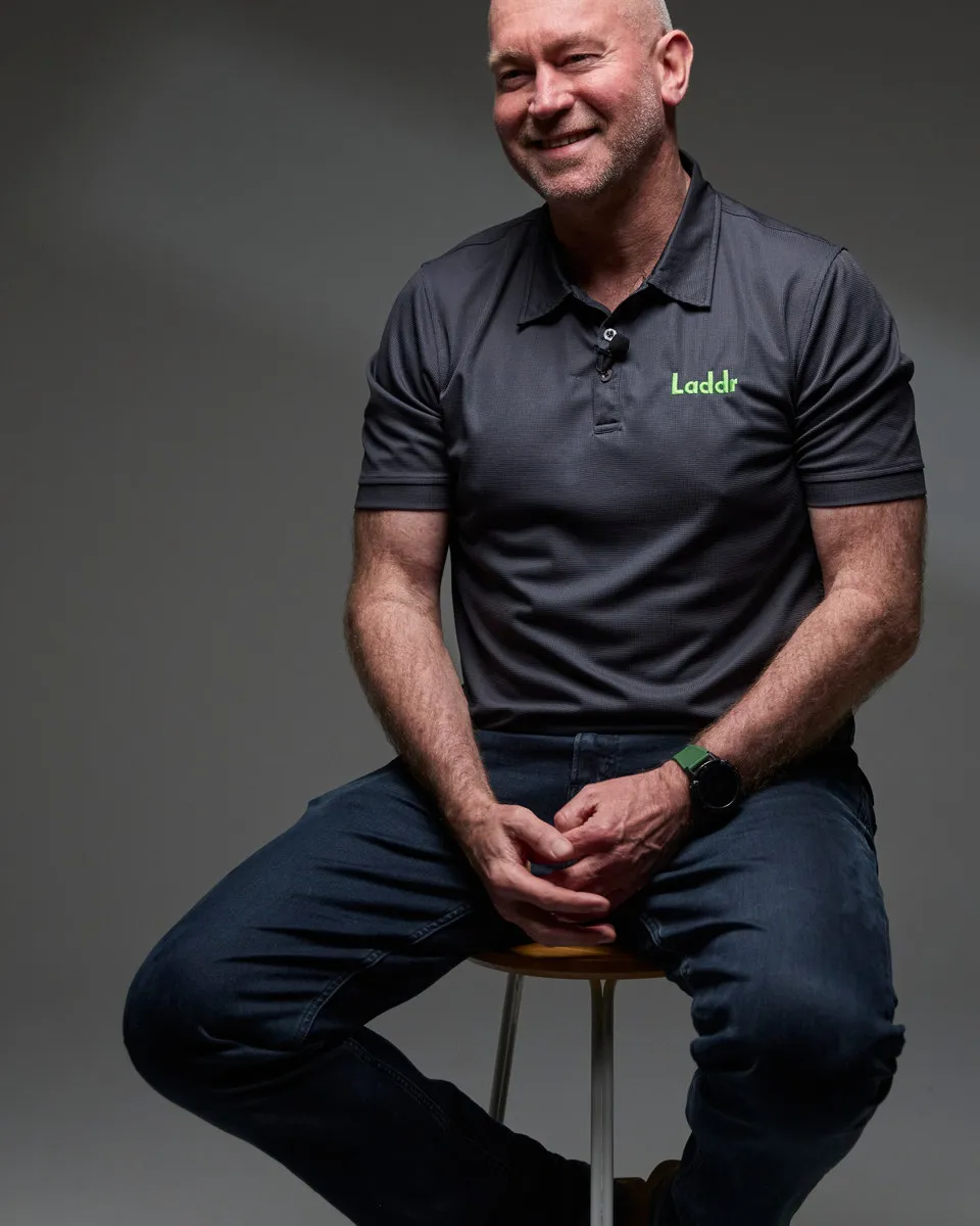 A smiling man in a Laddr polo shirt sitting on a stool against a grey backdrop