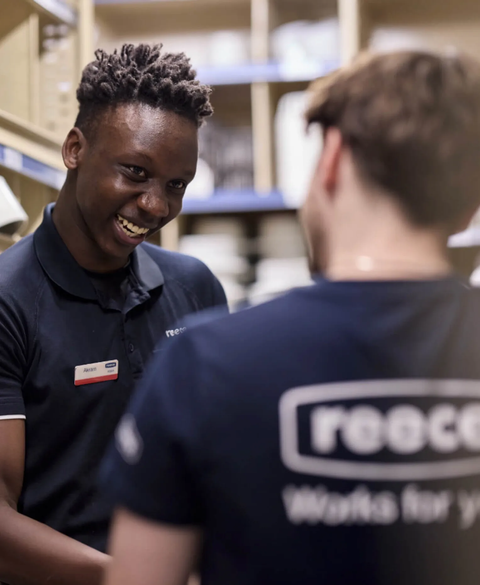 A smiling Reece team member in a navy polo talking with a colleague whose shirt reads “reece Works for you”, between shelves in a branch