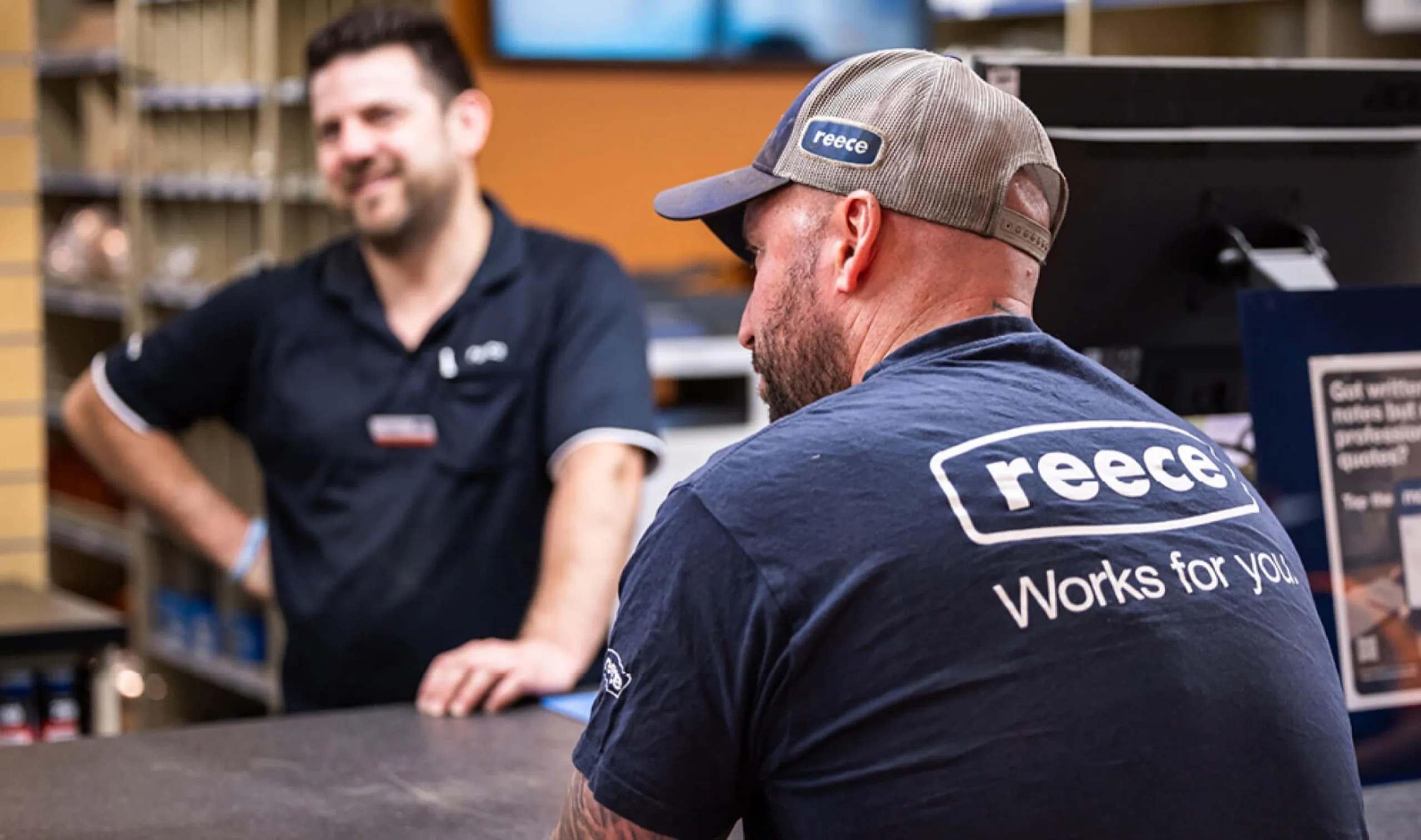 A tradie in a Reece cap and “Reece Works for you” t-shirt at a branch counter, a staff member smiling behind it