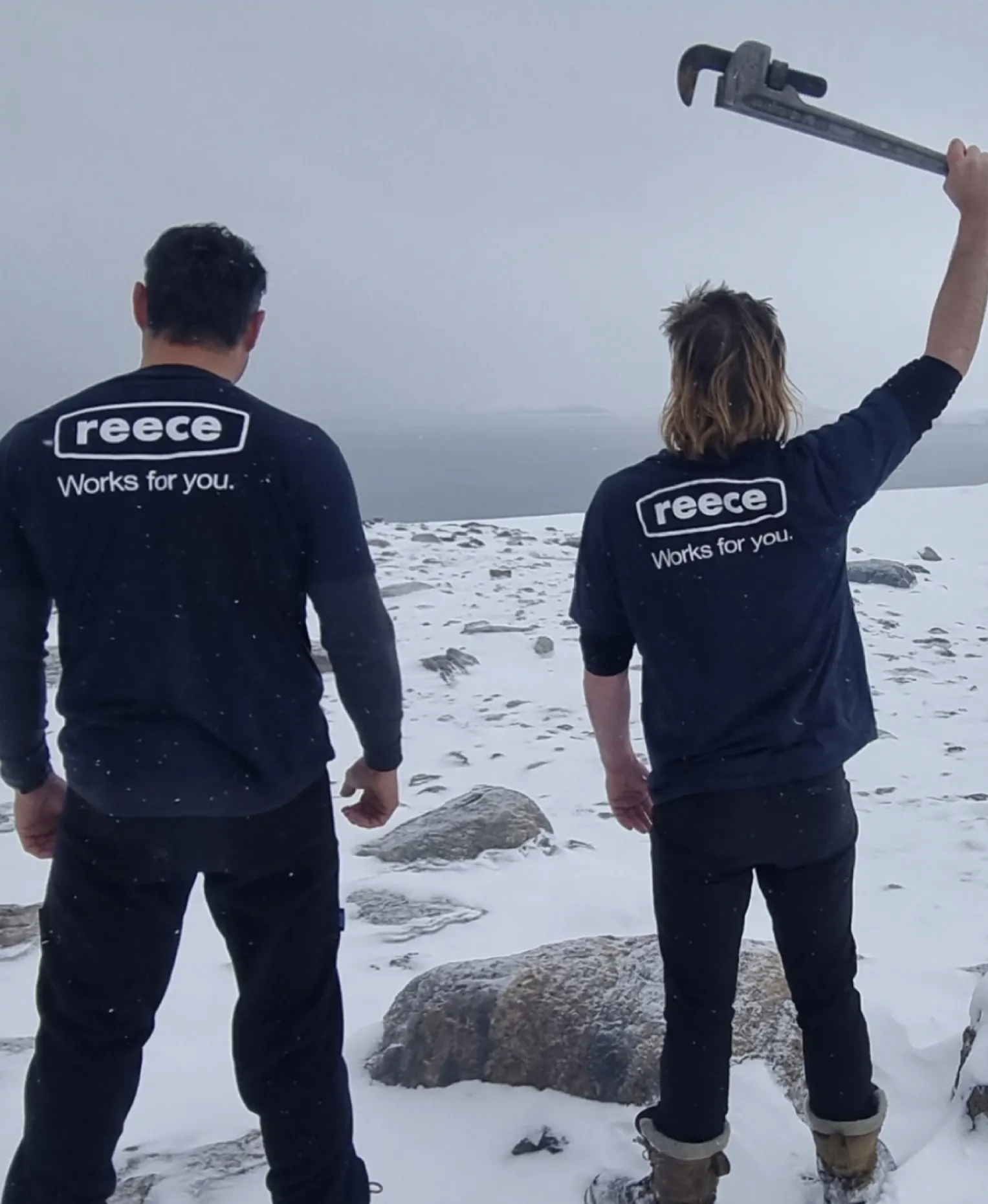 Two people in “Reece Works for you” t-shirts on a snowy mountaintop, one raising a pipe wrench