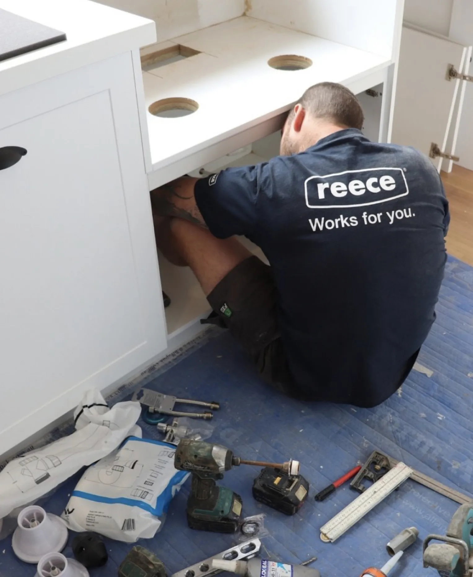 A plumber in a “Reece Works for you” t-shirt working under a kitchen sink, tools spread on the floor