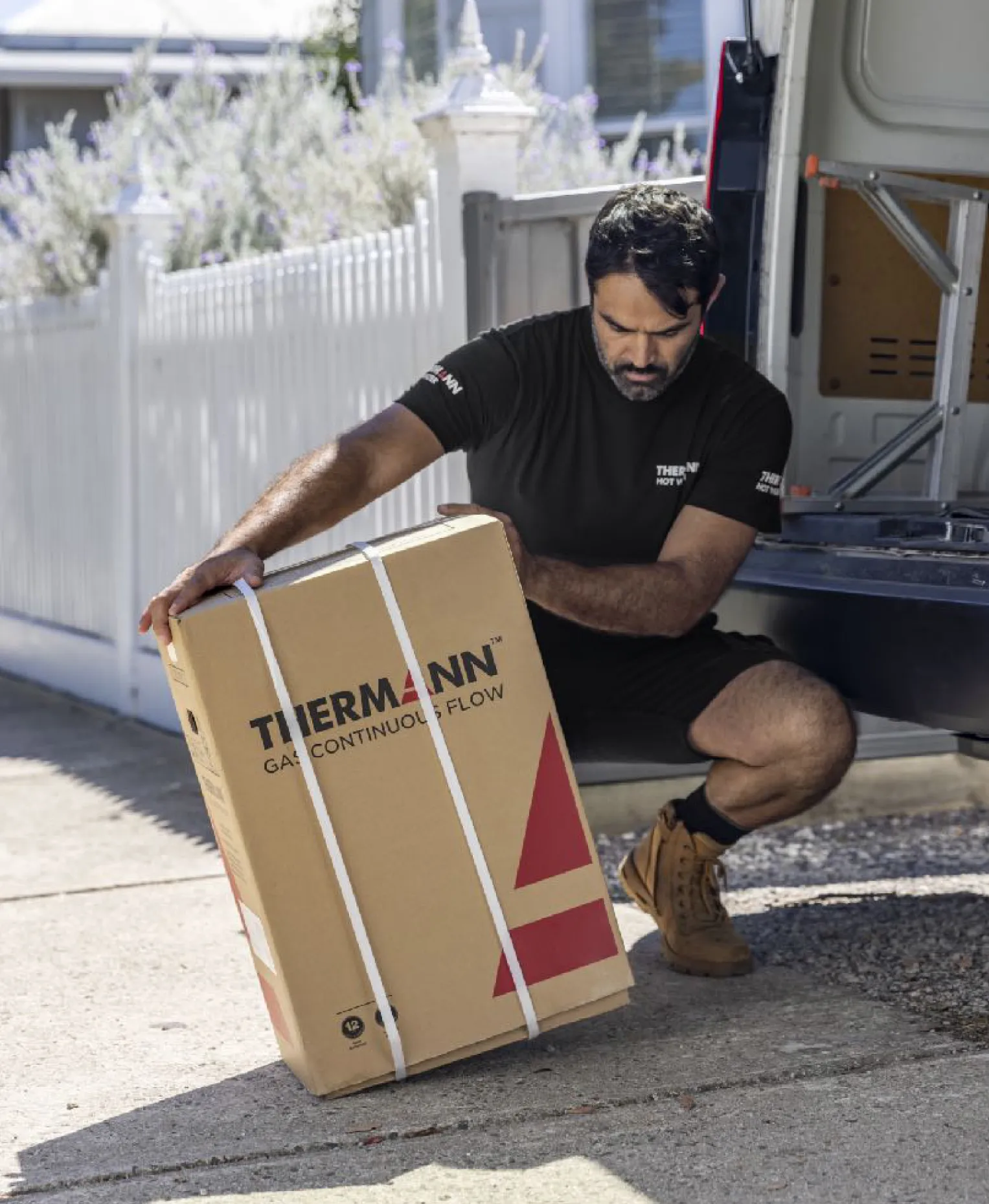 A tradie crouching on the footpath to lift a Thermann box marked “Gas continuous flow” out of the back of a van