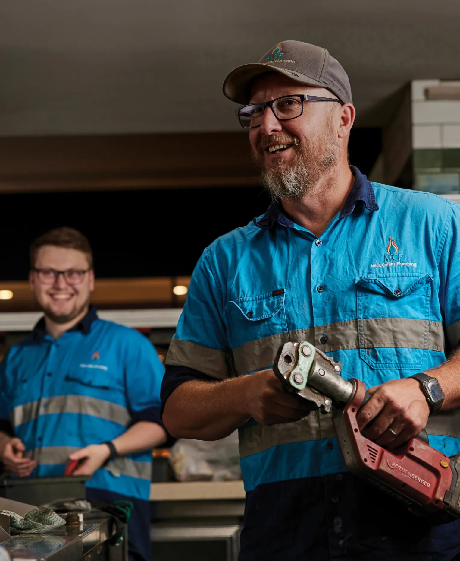 A smiling plumber in a cap and blue hi-vis shirt holding a red pipe-press tool in a commercial kitchen, a colleague laughing behind him