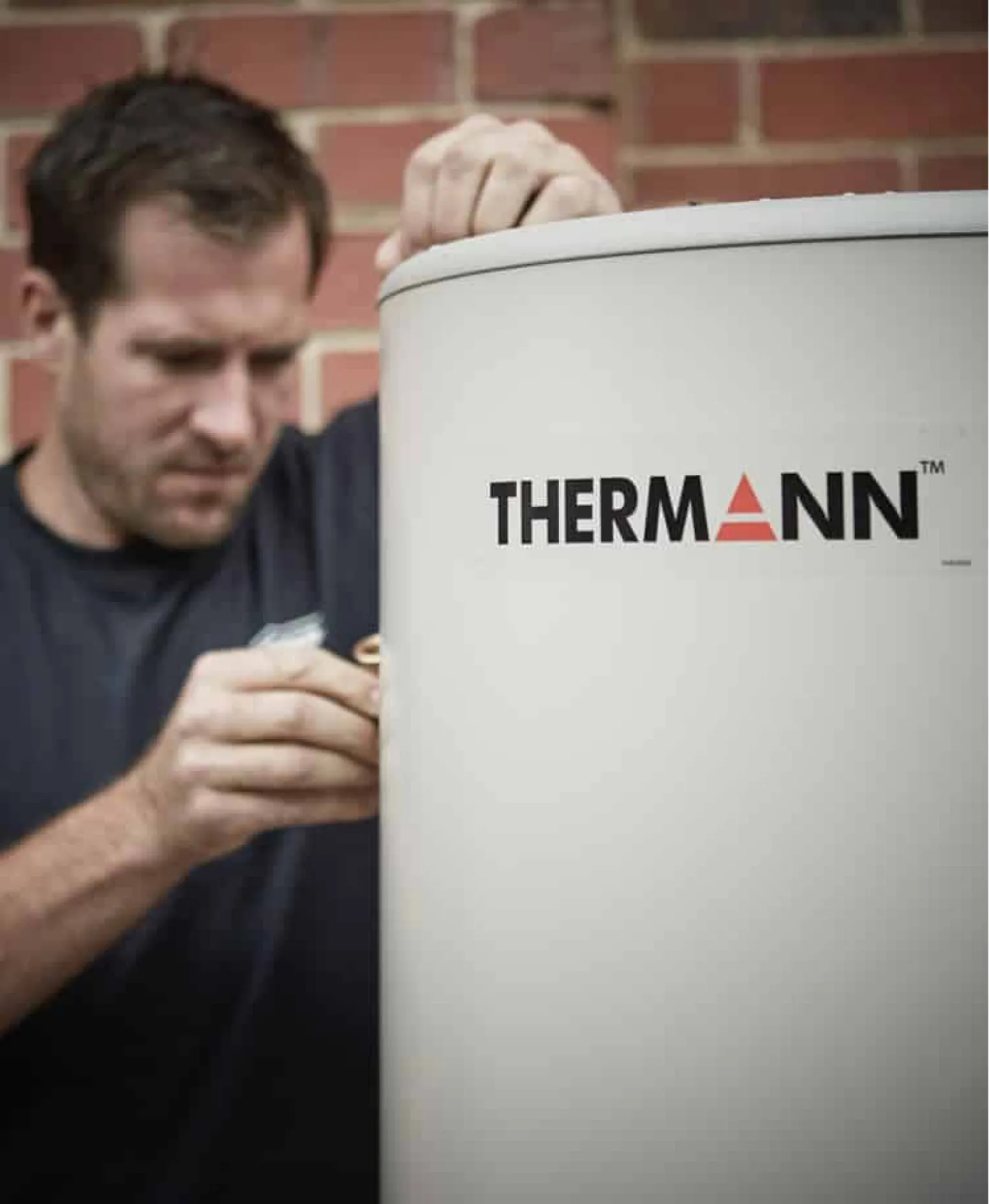 A tradie working on the top of a tall white Thermann hot water tank against a brick wall